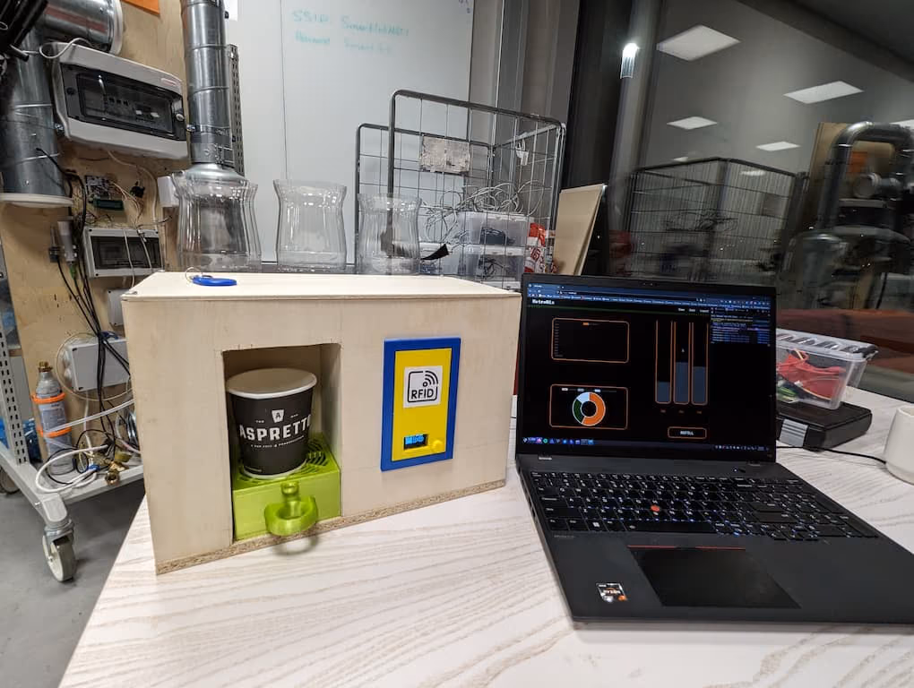 The finished PicoMix machine on a lab bench: a plywood box with a paper cup in the dispensing bay, an RFID reader on the front and three cut-open bottles on top